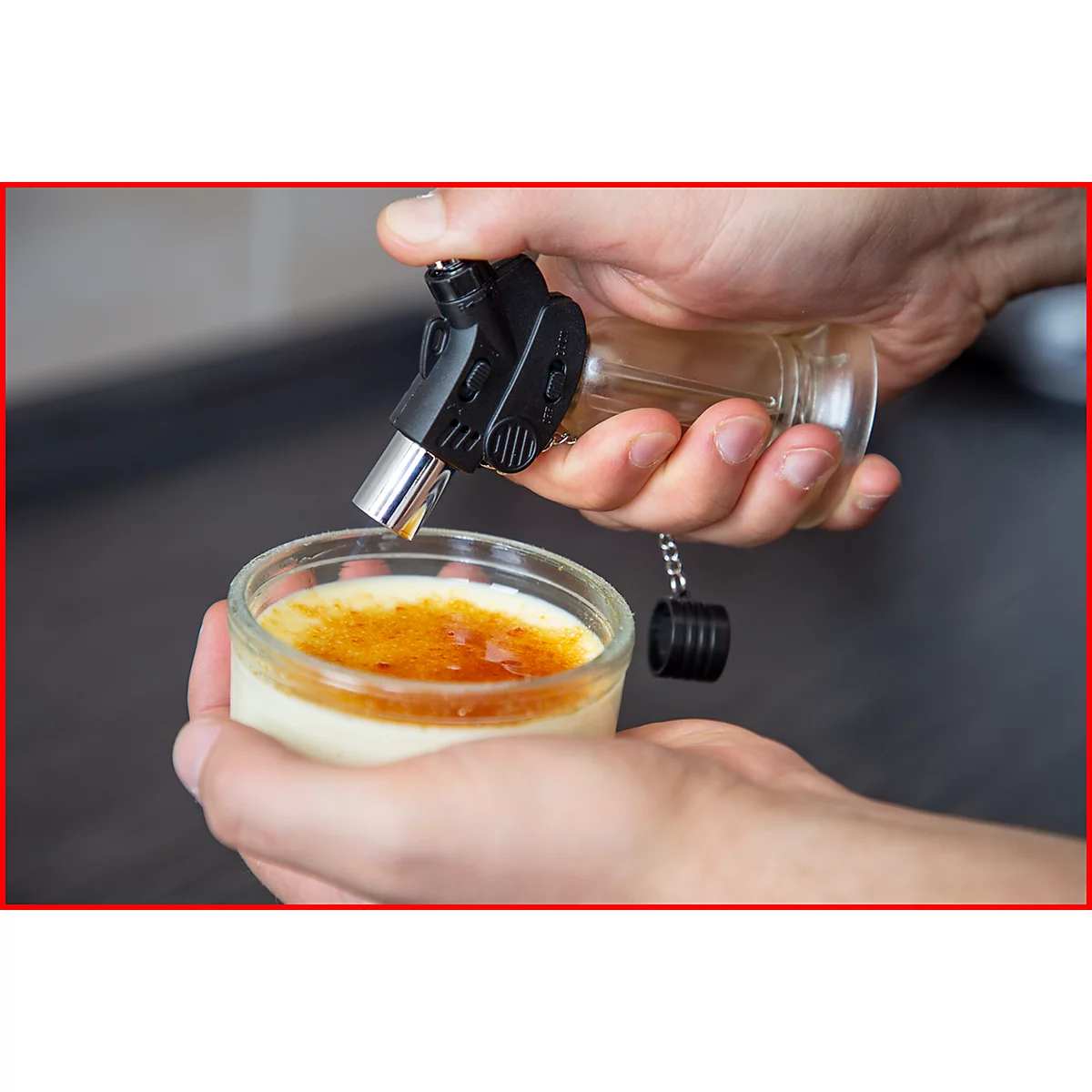 Person flambiert Crème brûlée mit einem Küchenbrenner. Das Dessert befindet sich in einem Glas.